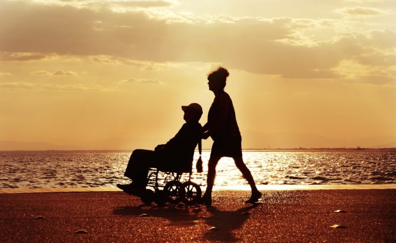 Una mujer conduce a un hombre con hemiparesia en silla de ruedas en la playa.