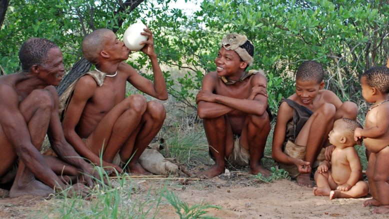 Jóvenes, ancianos, niños, mujeres y hombres negros en taparrabos, sentados en el suelo.